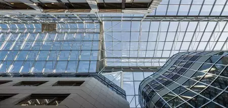 Kijk omhoog naar een architectonisch complex met een groot glazen dak en gevel, ondersteund door een metalen structuur. Rechts is een gebogen glazen gevel zichtbaar met een ruitvormig patroon. De constructie laat veel daglicht door en benadrukt modern en transparant ontwerp.