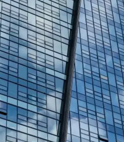 Hoekaanzicht van een moderne glazen wolkenkrabber tegen een deels bewolkte blauwe lucht. De gevel weerspiegelt de wolken, wat een patroon van licht en lucht op het gebouw creëert.