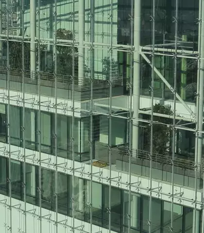 Uitzicht op moderne kantoorgebouwen in een stedelijke omgeving. Links een gebouw met een opvallende ruitvormige structuur, rechts een glazen toren met zichtbare stalen draagconstructie en beplante terrassen. Op de achtergrond een dichtbebouwde stad.
